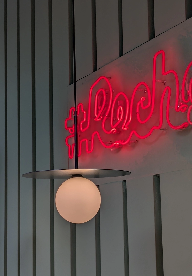 A close-up on modern interior design elements: a spherical matte pendant lamp with a flat metal disc hangs in the foreground, while a bright red neon sign glows on a slatted white wall behind it.