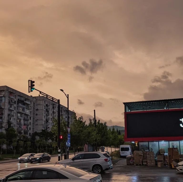 An urban street scene captured after rain, dominated by a dramatic, unfiltered orange and pink sky at dusk. On the left, a traffic light shows green above a Soviet-era apartment building, while on the right, a modern store building sits next to stacked boxes.