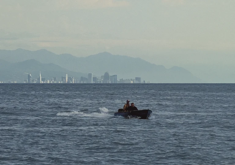 Two men in a small motorboat ride across the choppy Black Sea, creating a white wake. In the far distance, the entire city skyline of Batumi is visible, with a long range of hazy mountains rising behind it under a pale sky.