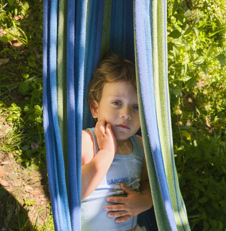 A young boy, Ted, lies in a blue and green striped hammock, looking directly up at the camera with a curious, contemplative expression as bright summer sun lights the green grass around him.