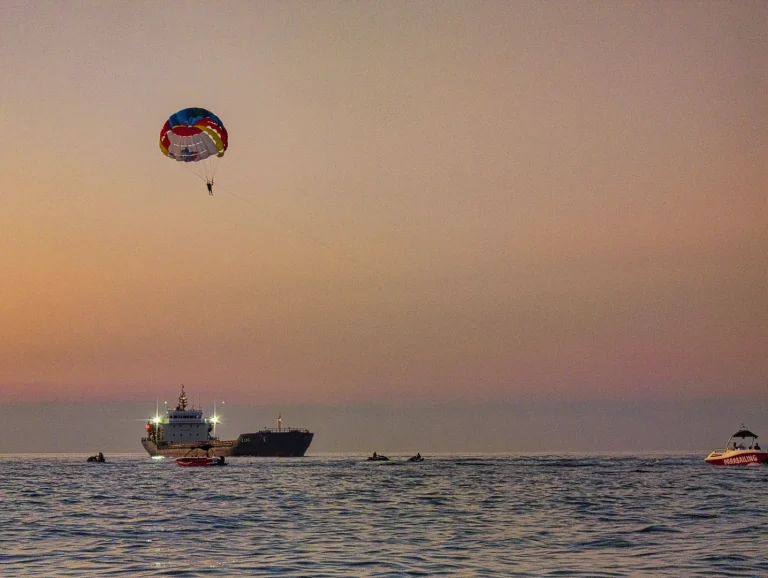 A dynamic Batumi seascape at sunset, featuring a person parasailing with a colorful parachute above a large cargo ship, with several jet skis and boats dotting the water against a hazy orange and purple sky.