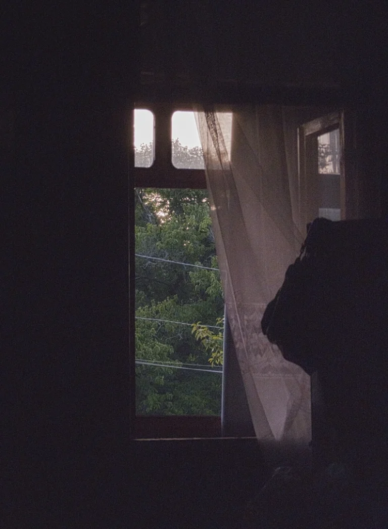 A view from inside a very dark room, looking out through a tall, narrow window onto lush green trees. The early morning sunlight illuminates the foliage and catches a sheer curtain hanging on the right, creating a stark contrast between the dim interior and the bright village morning outside.