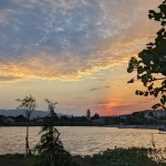 A horizontal landscape photo of a small, cozy lake in the suburbs of Tbilisi at sunset. The sky is a mix of purple, pink, and orange, with clouds reflected in the rippling water. The sun sets on the right horizon, and the far shore is lined with small houses and trees.
