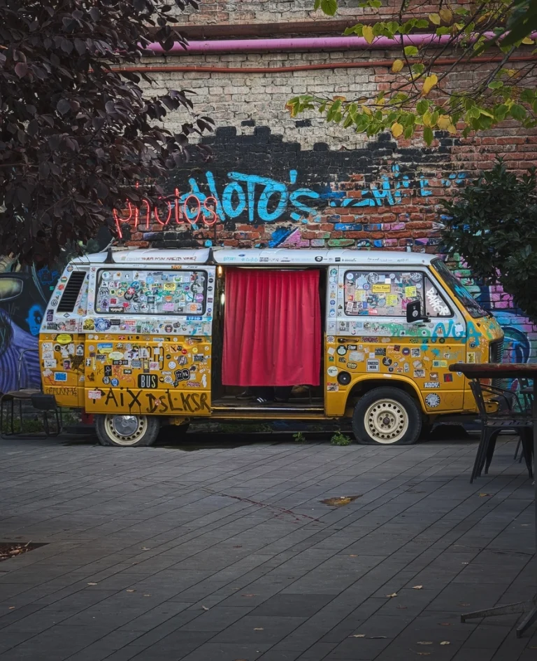A yellow vintage VW van, functioning as a photobooth, is parked in the Fabrika courtyard in Tbilisi. The van is covered in numerous stickers, and its open side door is draped with a red curtain. It sits in front of a colorful graffiti-covered brick wall.