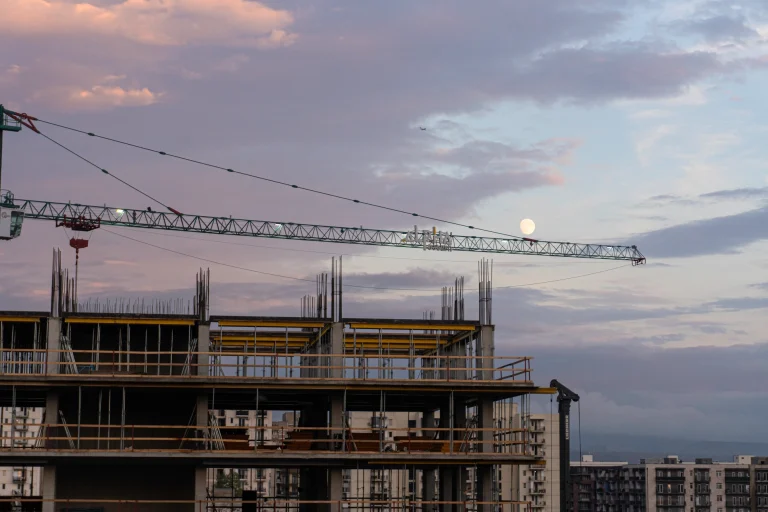 Night photo of a construction site crane in a Tbilisi suburb, with the full moon positioned as if rolling on its arm like a ball, and an airplane flying above. Captured by Georgian artist Zurab Kostava.