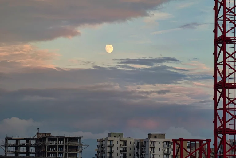 Photograph of a large, bright full moon rising above the silhouette of suburban buildings in Tbilisi, Georgia at night. Atmospheric cityscape photography by Georgian artist Zurab Kostava.