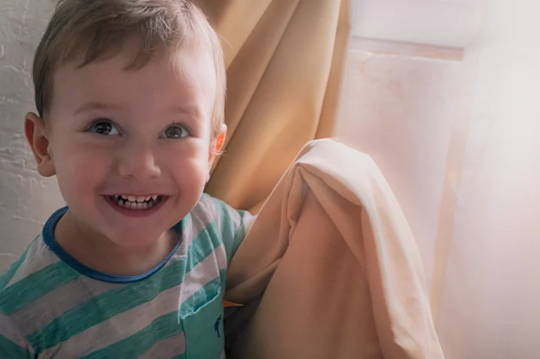 Young Ted Kostava, son of Georgian artist Zurab Kostava, playfully hiding behind a curtain illuminated by morning sunbeams around age two. Captures childhood innocence and joy. Photo by his father.