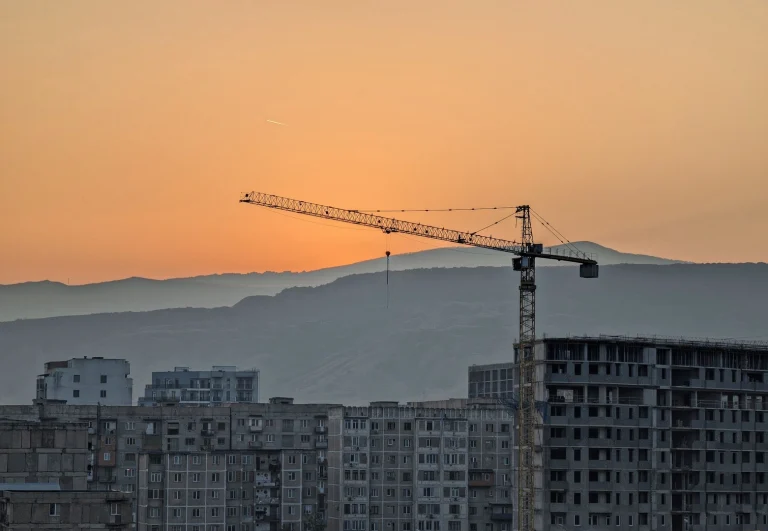 A tall construction crane stands silhouetted against a vibrant orange sunset. In the foreground are Soviet-era apartment buildings, with hazy mountains visible in the far distance behind the crane.