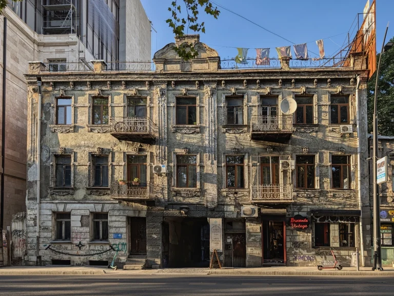 The weathered, ornate facade of an old, multi-story building in Tbilisi, with crumbling plaster and balconies. Modern air conditioners and a satellite dish are attached, and laundry hangs on the roof. A modern glass building is visible right next to it.