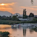 A vertical photo of a lake at sunset. A small white pier with people on it extends into the water, casting clear reflections. Sunbeams shine over distant mountains, illuminating houses and an electricity pylon on the shore, which is lined with tall reeds. A willow branch hangs in the top right corner.