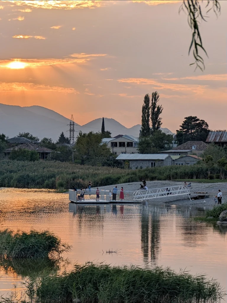 A vertical photo of a lake at sunset. A small white pier with people on it extends into the water, casting clear reflections. Sunbeams shine over distant mountains, illuminating houses and an electricity pylon on the shore, which is lined with tall reeds. A willow branch hangs in the top right corner.