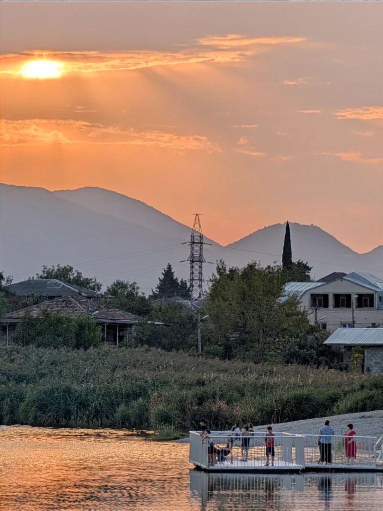 Several people stand on a small, white floating pier on a lake at sunset. The orange sky and sun cast dramatic rays over hazy mountains in the background, with the reflections glowing on the water.